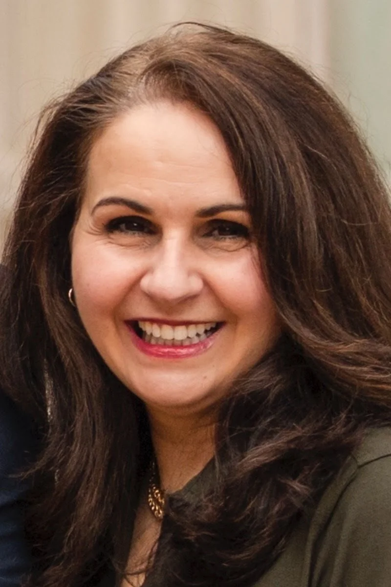 A woman with dark hair smiling, wearing a tan coat and standing indoors.
