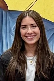 A young woman with long brown hair smiling, wearing a black jacket, in front of a yellow and blue background.