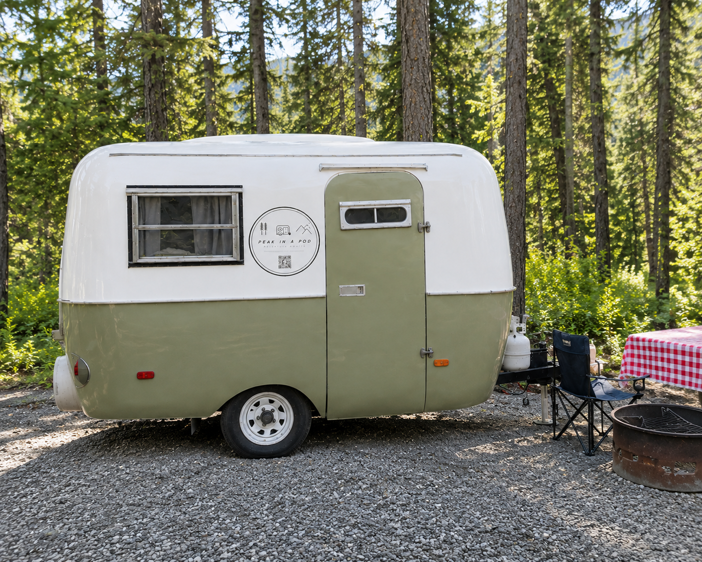 A vintage green and white camper trailer parked in a forested area with tall trees. There is a small window with curtains, a logo on the side that reads 'Peak in a Pod,' and an outdoor setup including a foldable chair, a table with a red and white checkered tablecloth, a propane tank, and a fire pit.