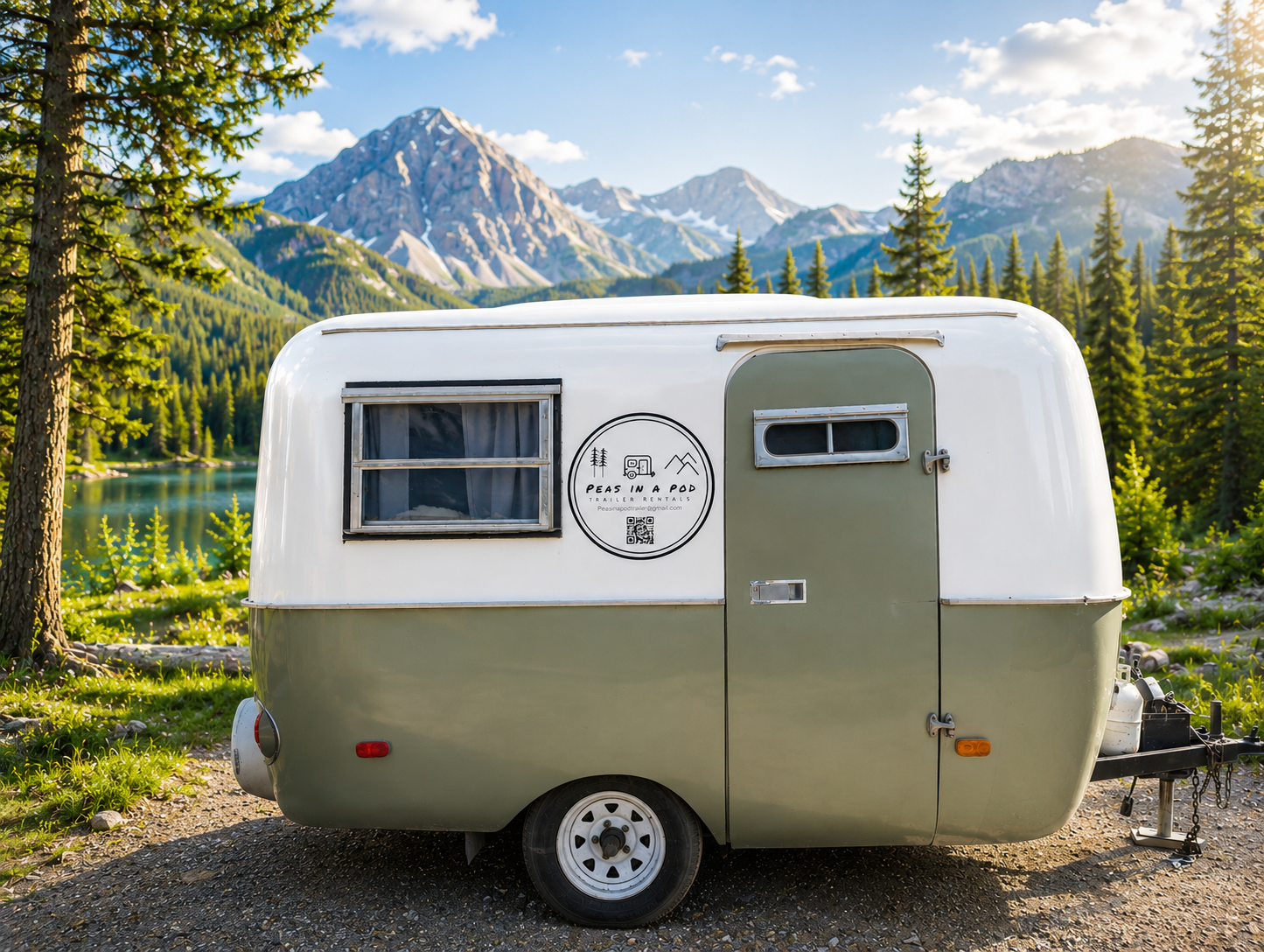 Small vintage camper trailer parked outdoors in front of a scenic mountain and forest landscape, with a lake nearby.