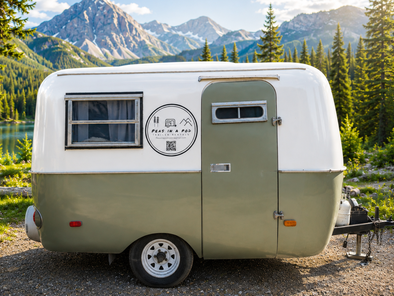 A vintage camper trailer in light green and white, parked on a gravel ground with a scenic mountain and forest background, featuring a lake and tall pine trees.