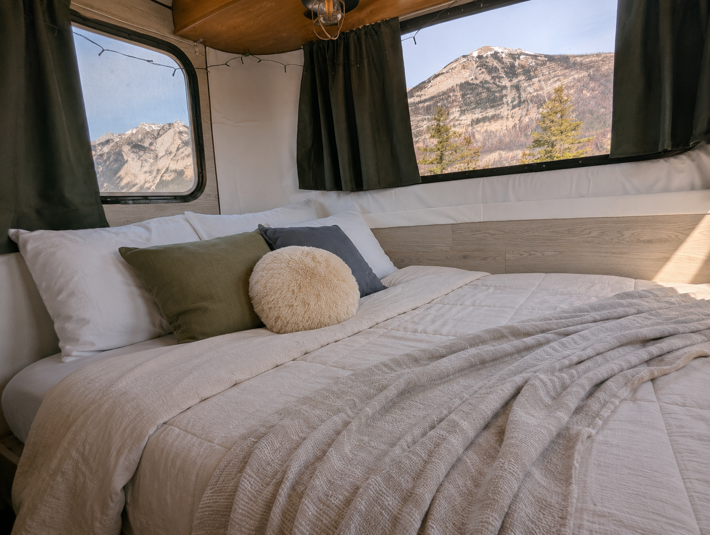 A cozy bed inside a tiny house camper with mountain and pine tree views through large windows, decorated with pillows and a cream-colored blanket.