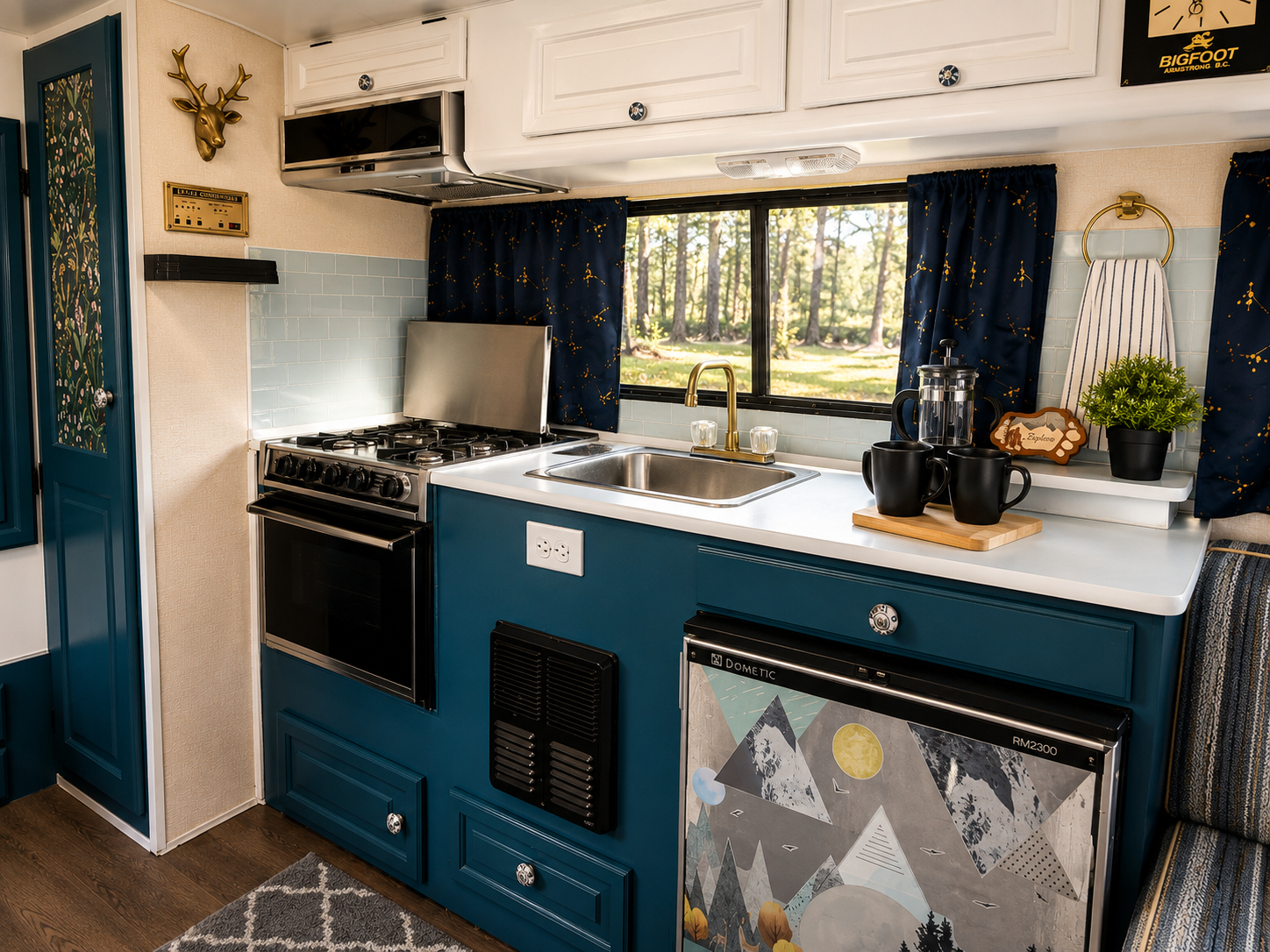 A cozy kitchen with blue cabinets, a white countertop, and a window overlooking a wooded area. The kitchen features a black stove with a matching hood, a stainless steel sink with a gold faucet, and decorative items including black mugs on a wooden t