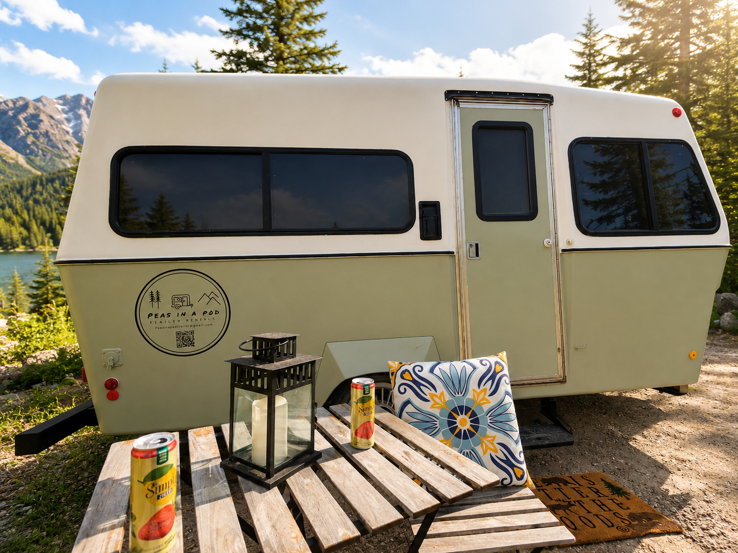 A camper trailer parked outdoors near a lake and mountains, with a small wooden table in front holding two cans of juice, a lantern, and a decorative pillow.