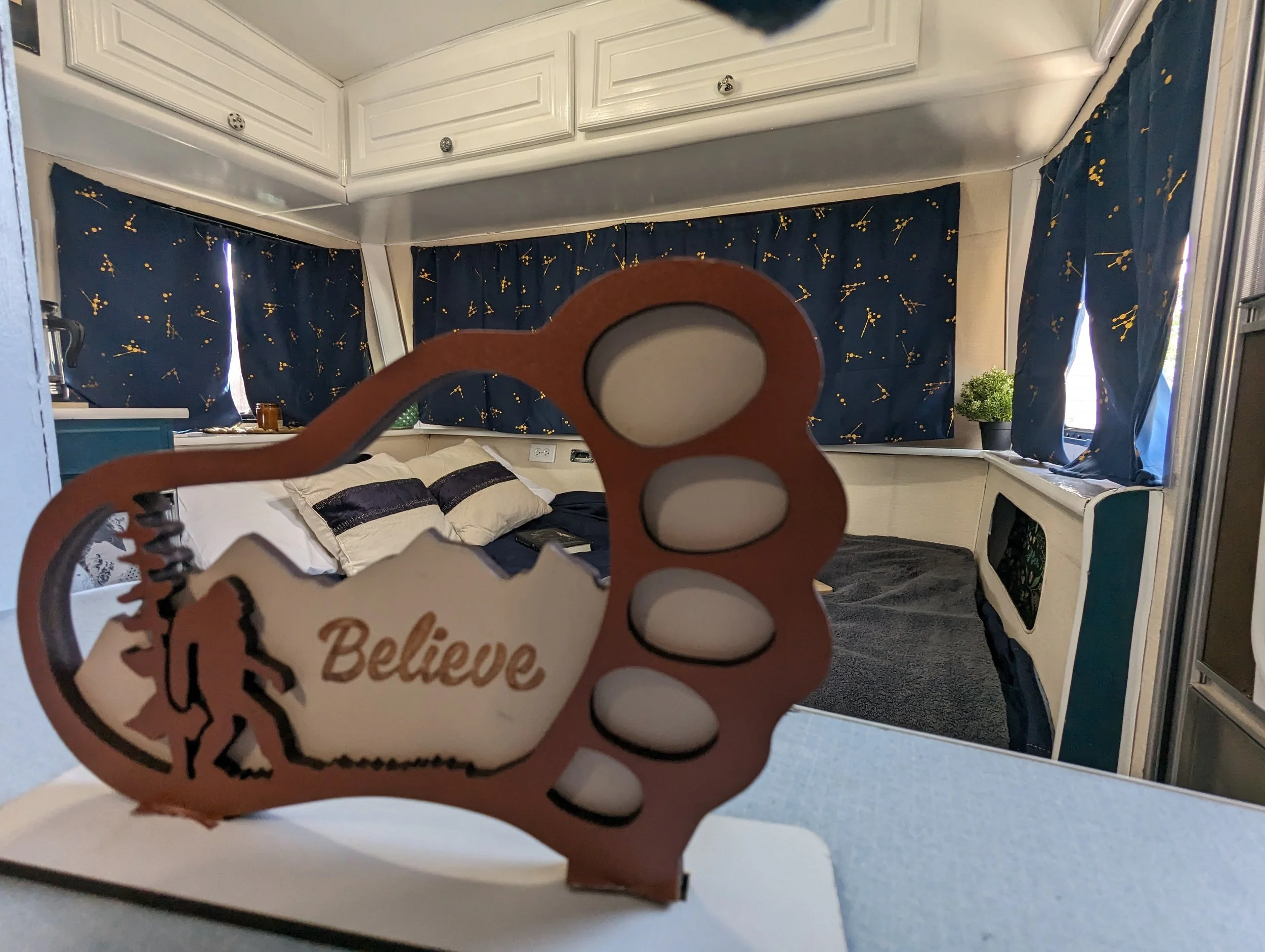 Room with white cabinets, dark curtains with a yellow floral pattern, a bed with pillows, a small green potted plant, and a mirror with a wooden frame that says "Believe".