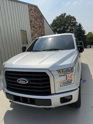 White Ford pickup truck parked outdoors on a concrete surface near a building with metal siding and brick wall, under a blue sky with some clouds.