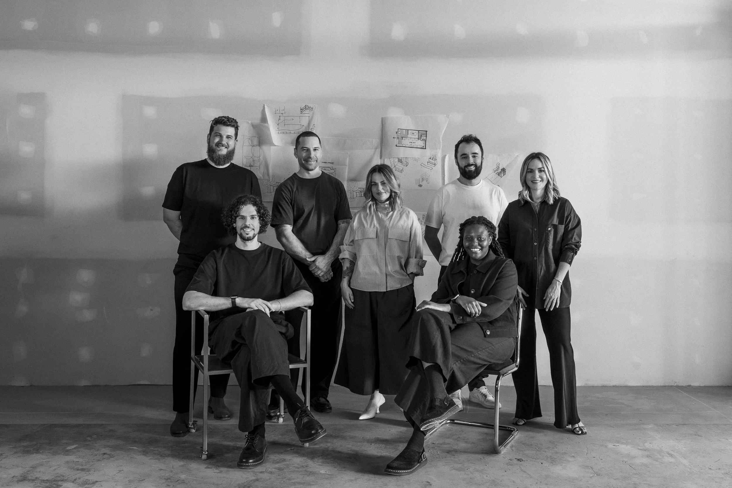 A black and white photo of a diverse group of seven people, standing and sitting, inside an industrial-style space with sketches and plans on the wall behind them.