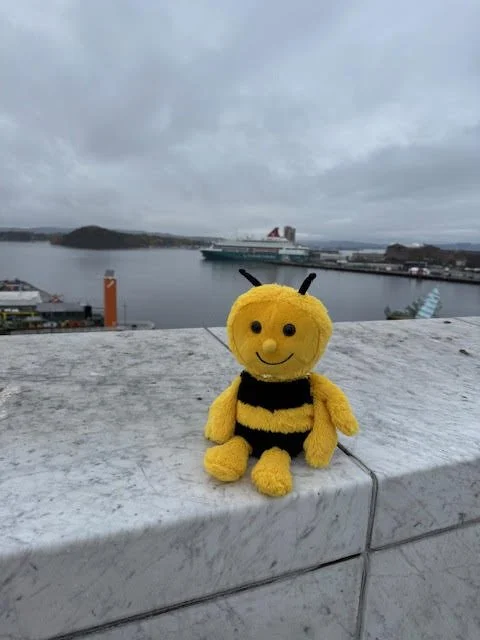 A yellow plush bee toy with black stripes and black antennae sitting on a white stone ledge overlooking a harbor with ships, water, and overcast sky.