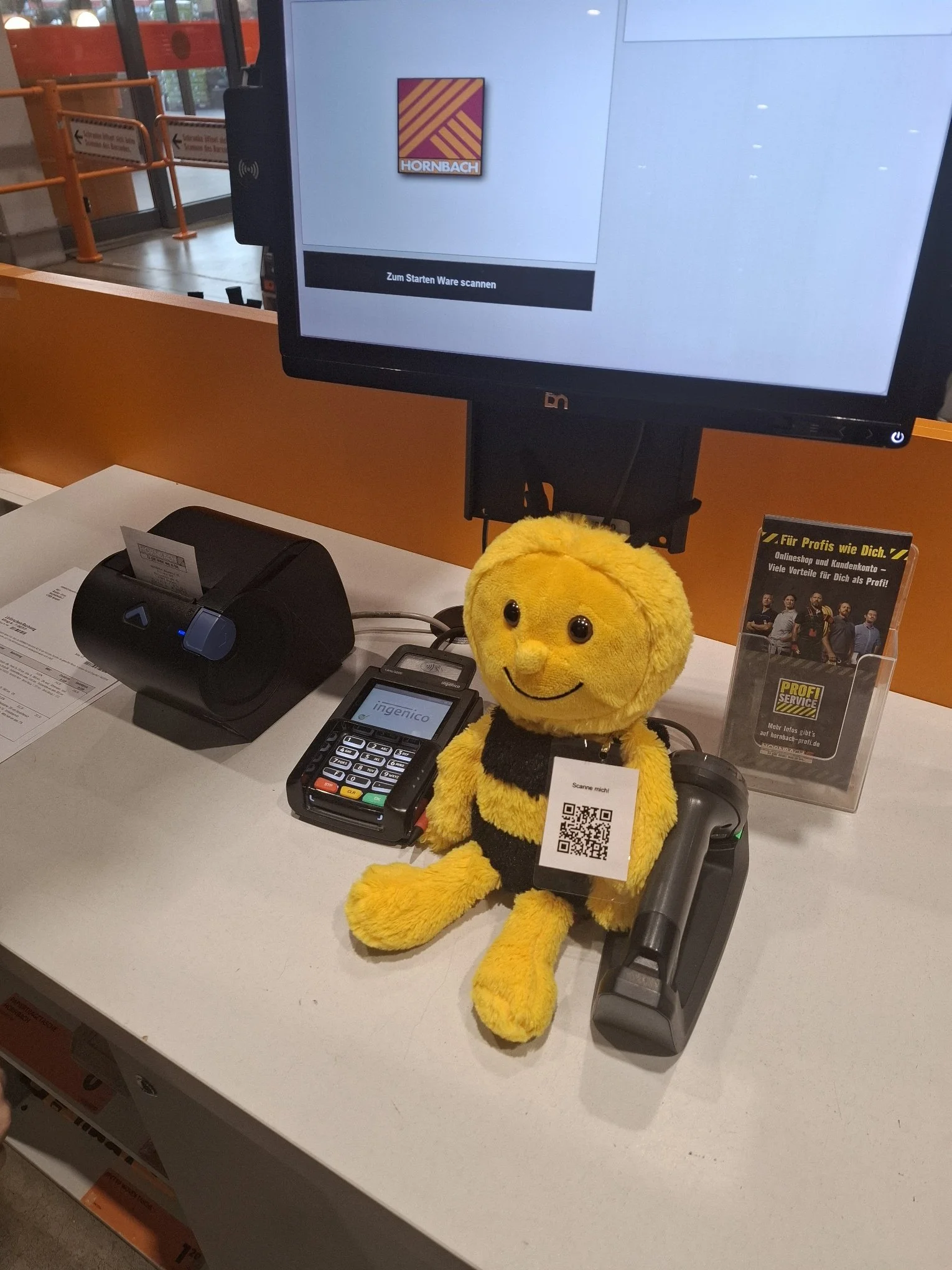 Ein Kuscheltier in Form einer Biene mit einem QR-Code am Hals, das vor einem Kassensystem und einem Monitor in einem Geschäft steht.
