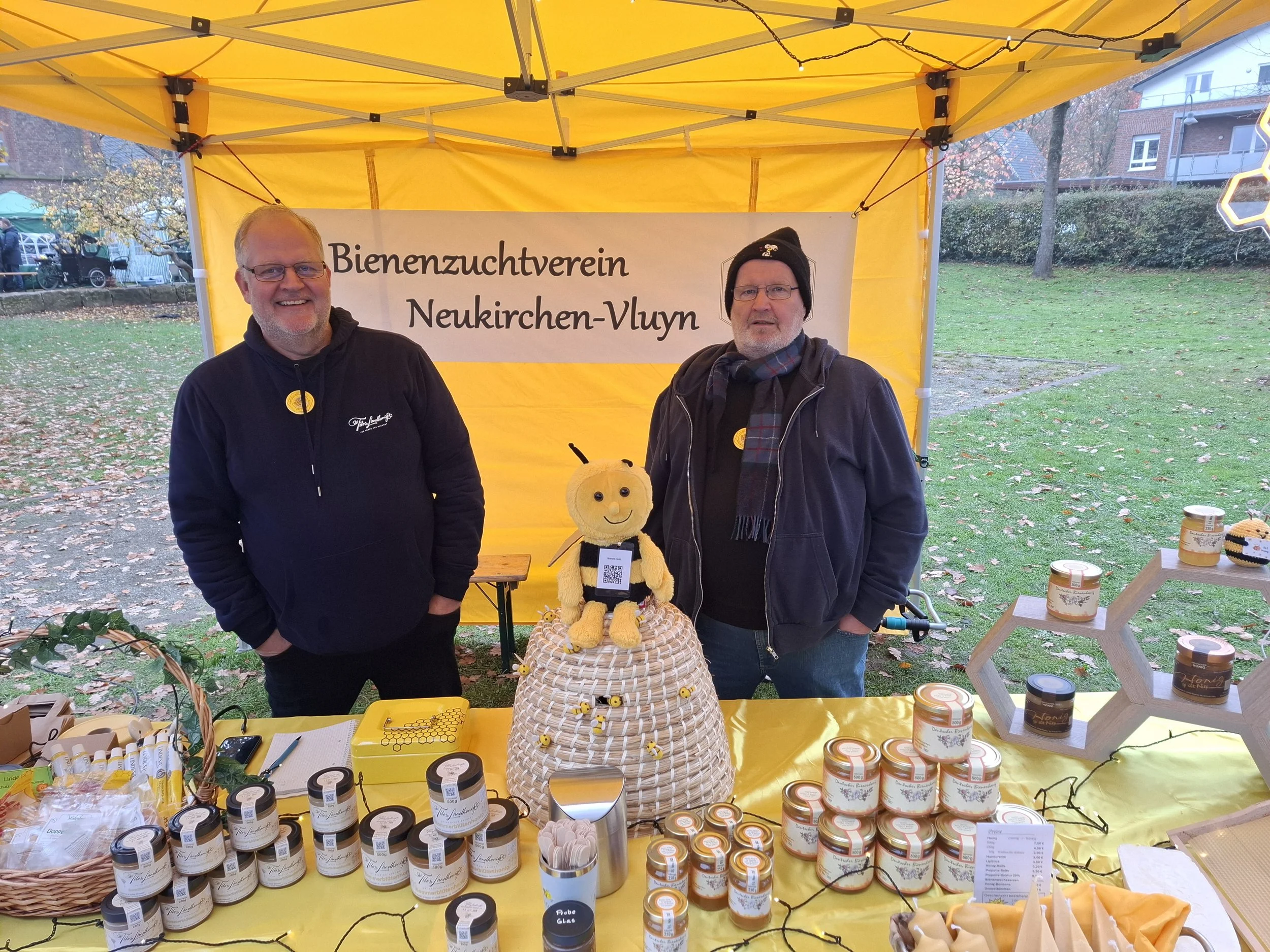 Zwei Männer stehen an einem Verkaufsstand der Bienenzuchtverein Neukirchen-Vluyn, vor einem gelben Zelt. Der Verkaufsstand hat Honiggläser, eine Honigwabe und eine Plüschbiene. Im Hintergrund sind Bäume und Häuser zu sehen.