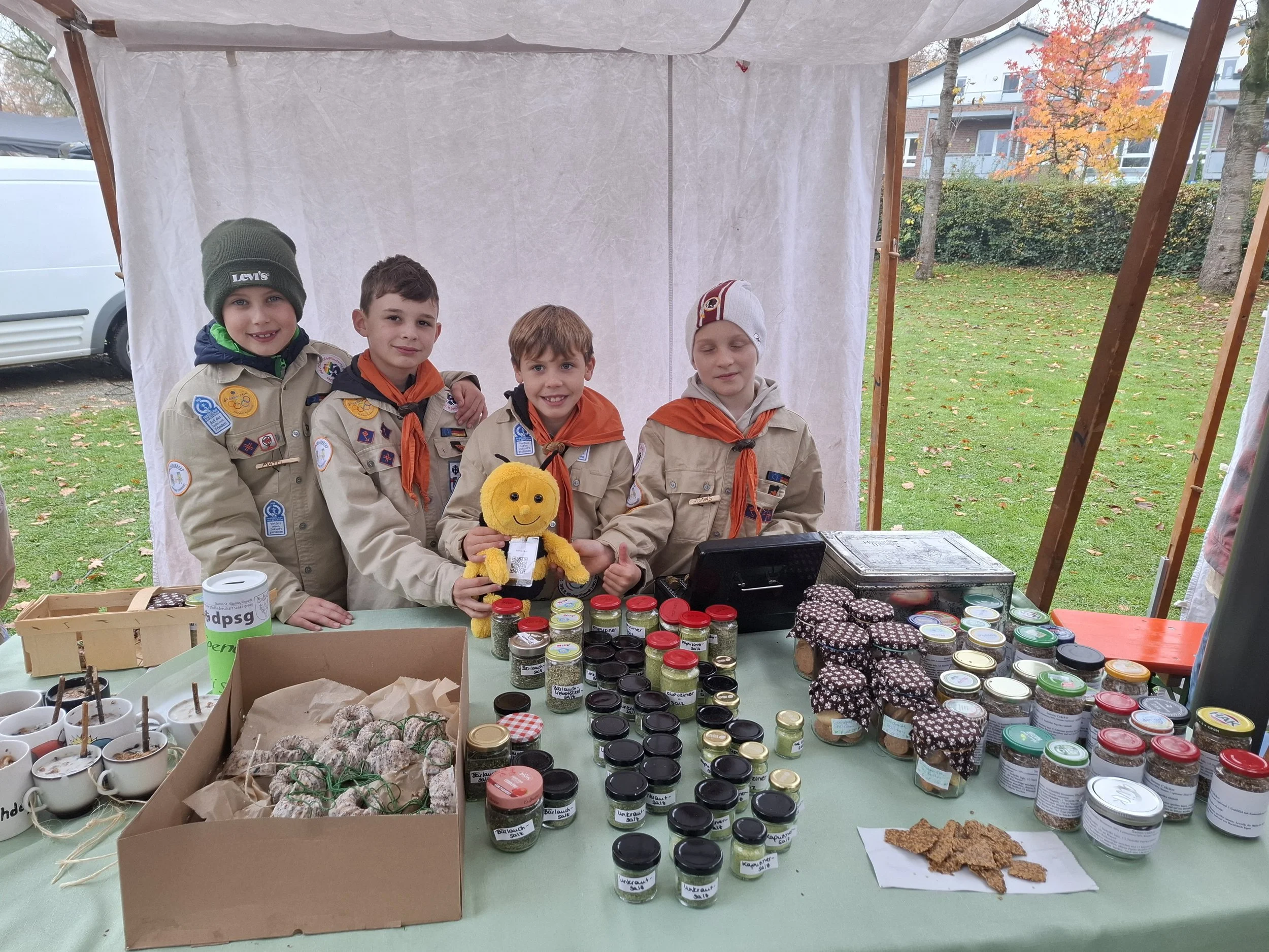 Vier Jungen in Pfadfinderuniformen stehen hinter einem Verkaufstisch mit Marmeladen, Keksen und Kaffee. Einer der Jungen hält einen gelben Plüschhund. Es ist ein gemischtes Herbstwetter mit fallen Laub im Hintergrund.