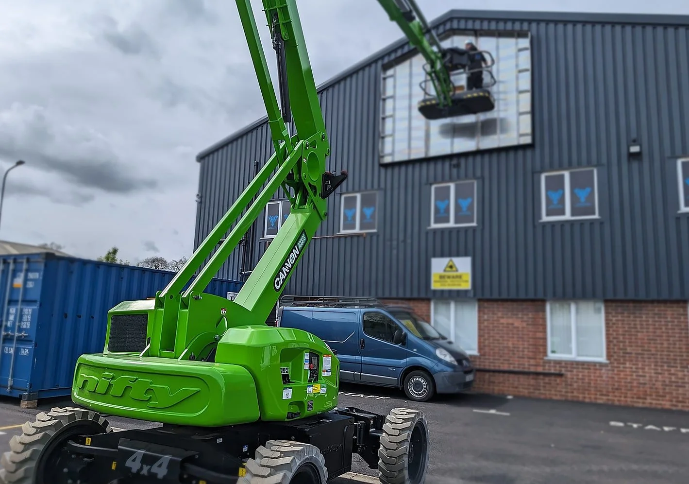 Green Nifty Boom lift extending toward the upper floor of a dark gray industrial building, with a blue van parked nearby and a large blue shipping container to the side.