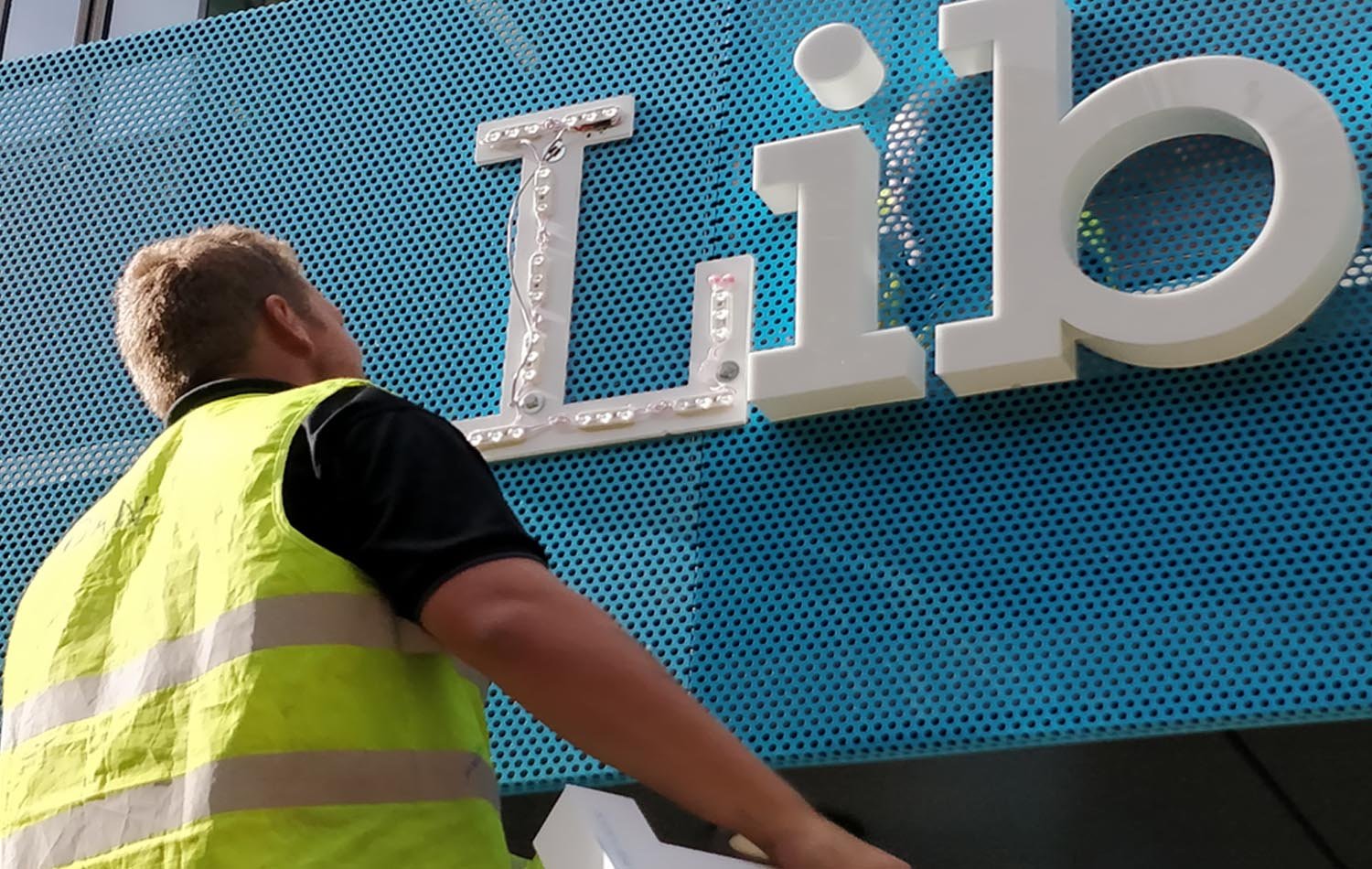Person in a yellow safety vest installing or maintaining a large white sign on a blue perforated wall. The sign spells out 'T.io' with decorative elements.