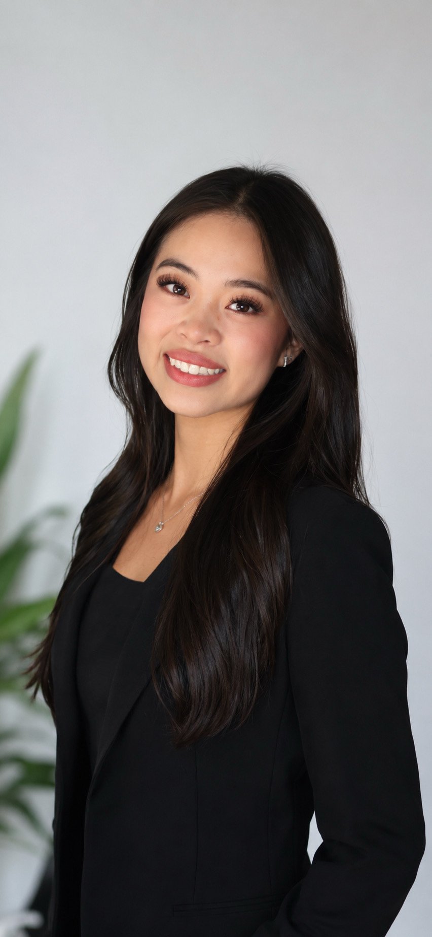 Portrait of a young woman with long dark hair wearing a black top and a necklace, posed against a gray textured background.