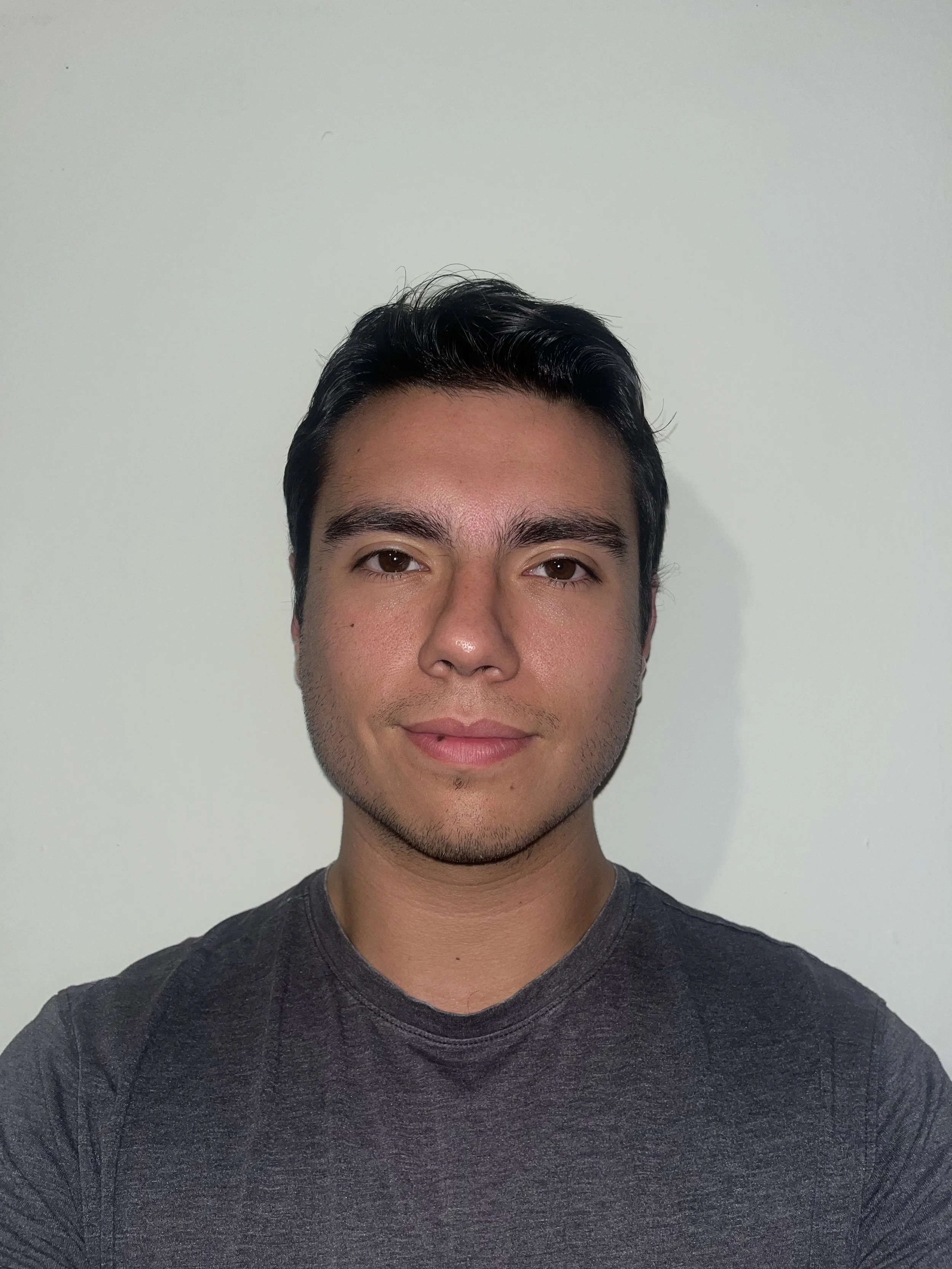 Portrait of a young man with dark hair in front of a plain light-colored wall.