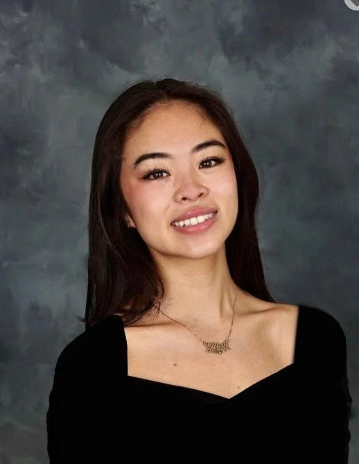 Portrait of a young woman with long dark hair wearing a black top and a necklace, posed against a gray textured background.