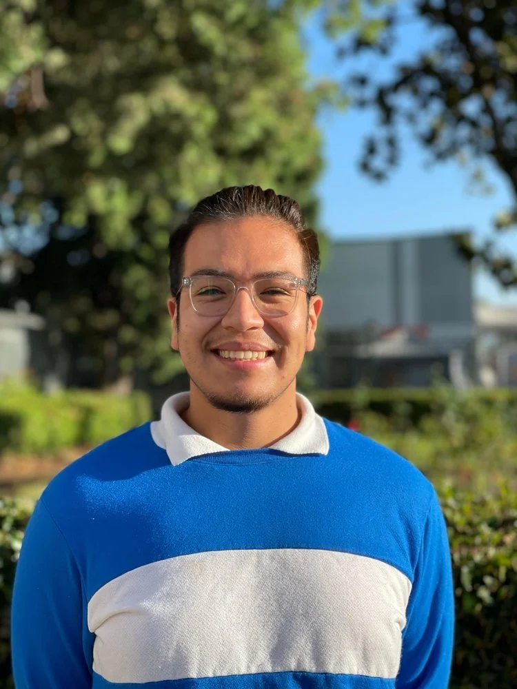A young man with glasses, dark hair, and a goatee smiling outdoors on a sunny day, wearing a blue and white striped shirt with a background of trees and a building.