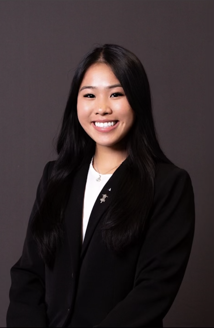 A young Asian woman with long black hair smiling, wearing a black blazer and a white top, standing against a dark grey background.