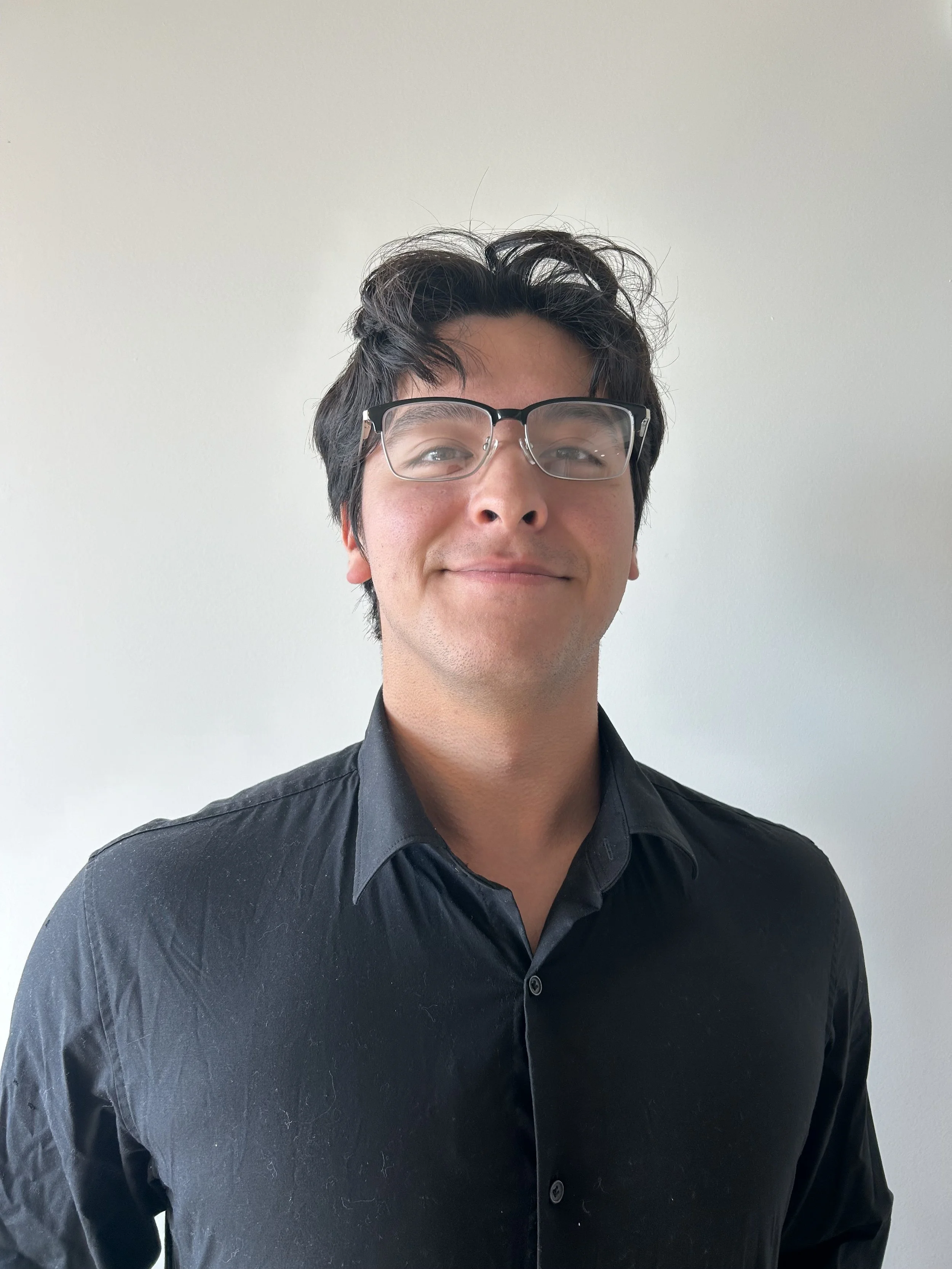 A young man with dark hair, glasses, and a slight smile, wearing a black shirt, standing against a plain white wall.