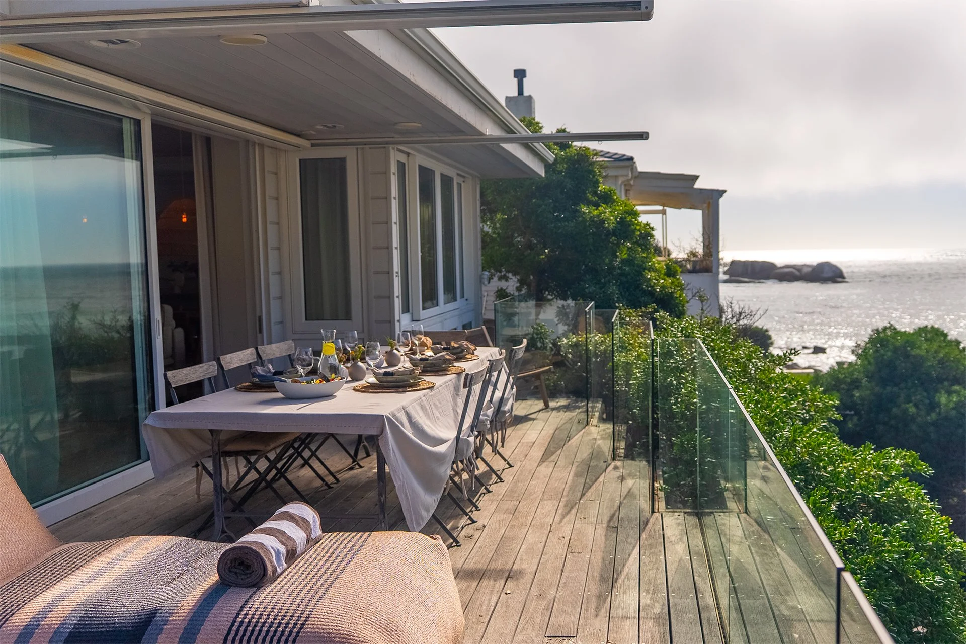 Long glass-walled beachfront deck with sunbeds and outdoor dining table for eight guests with braai, with BBQ area behind the kitchen. Direct access to beach down 40 steps. Make an enquiry for our Cape Town luxury villa.