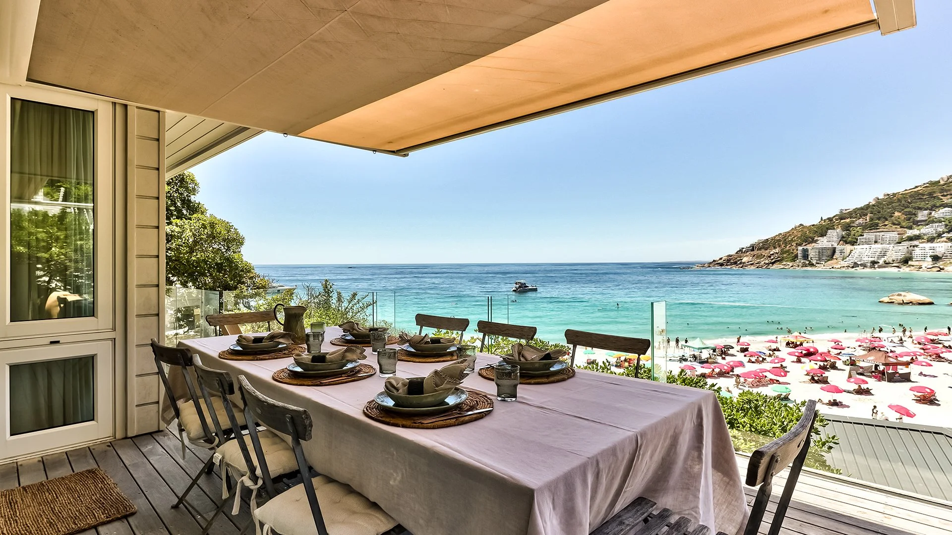 Long glass-walled beachfront deck with sunbeds and outdoor dining table for eight guests, with braai and BBQ area behind the kitchen. Secure your booking now for a Cape Town getaway. 
