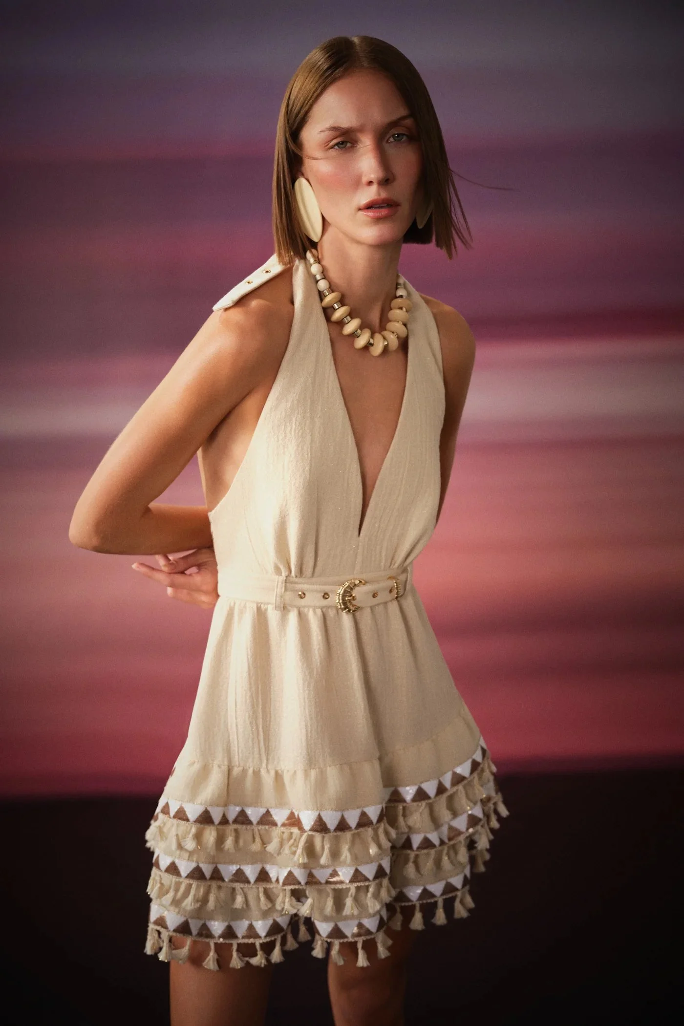 A woman wearing a beige sleeveless dress with a V neckline, decorated with tassels and geometric pattern at the hem, paired with large earrings, a chunky shell necklace, and a belt with a gold buckle, standing against a blurred sunset background.