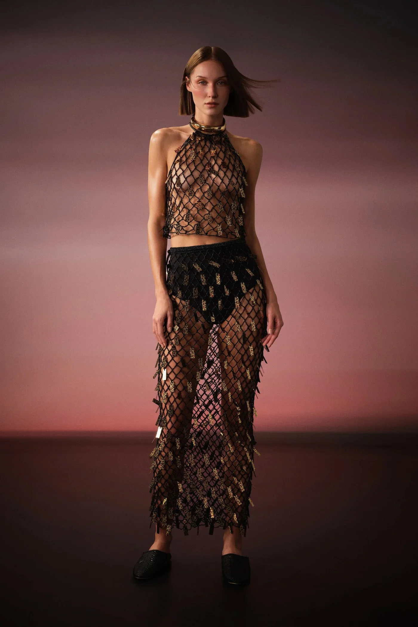 Fashion model wearing black and gold fishnet outfit with jewelry, standing on a dark runway with pink and purple gradient background.