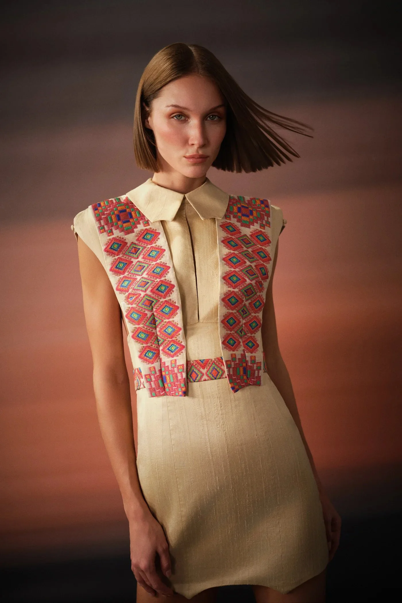 A woman with shoulder-length hair and a neutral expression wearing a beige dress with embroidered colorful geometric patterns on the shoulders and chest, standing against a blurred gradient background.