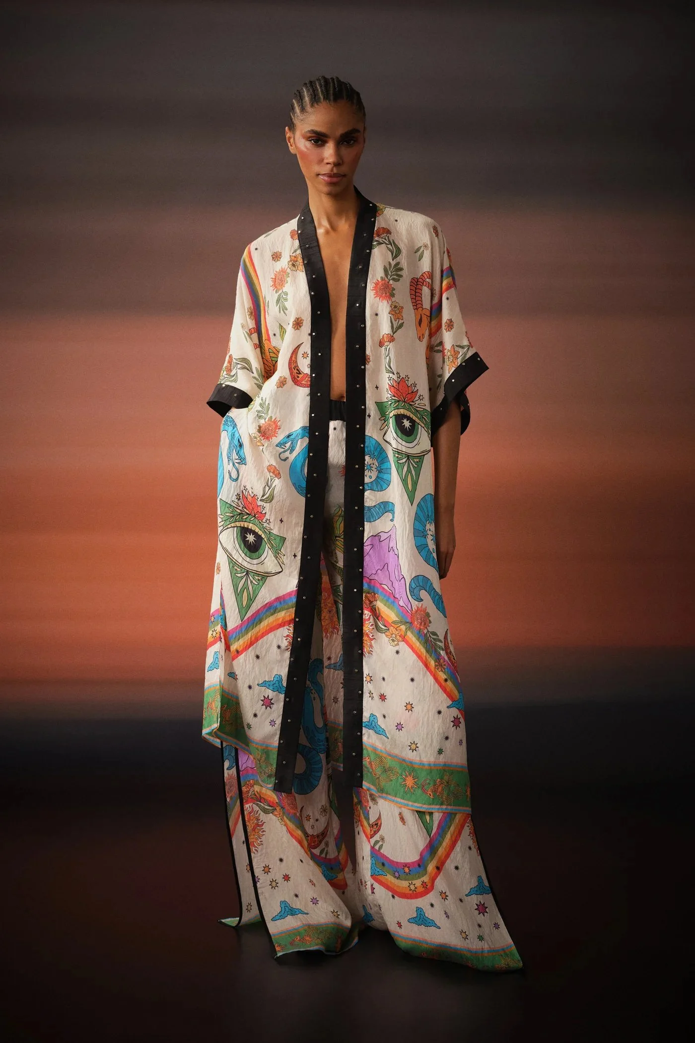 Model wearing a long, colorful, paisley-printed kimono-style robe with black trim and wide-leg matching pants, standing against a sunset backdrop.