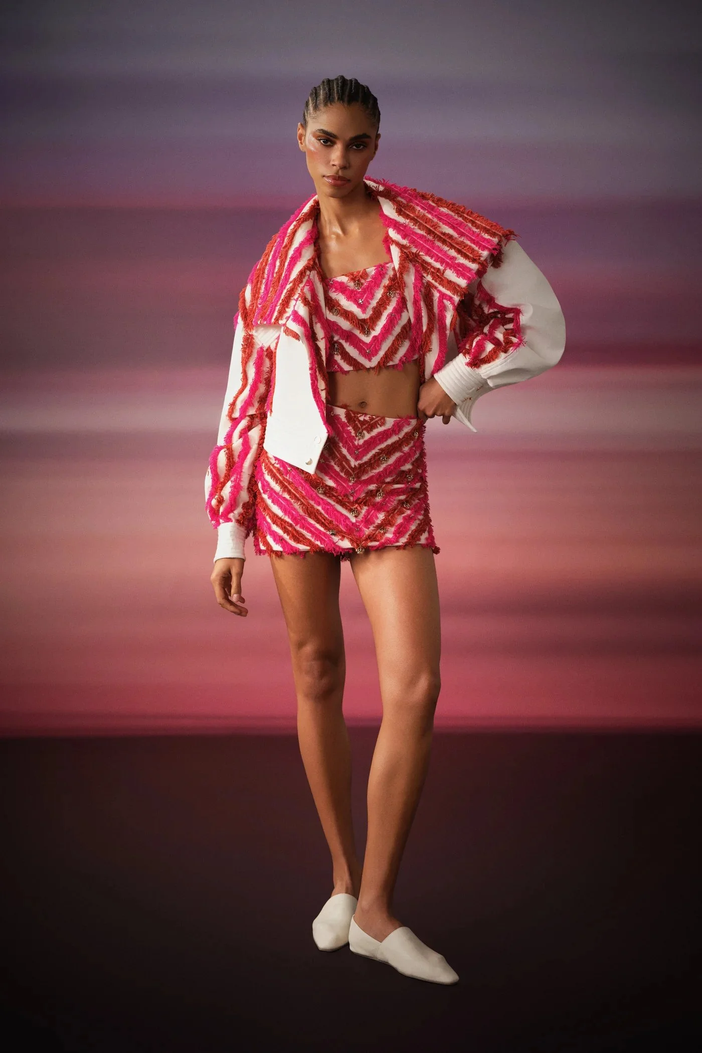A woman models colorful, textured pink and white clothing against a sunset background.