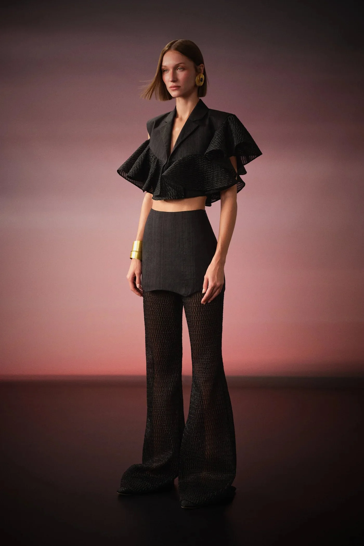 A woman wearing a black ruffled crop top, high-waisted wide-leg pants, and gold accessories, standing against a gradient background.