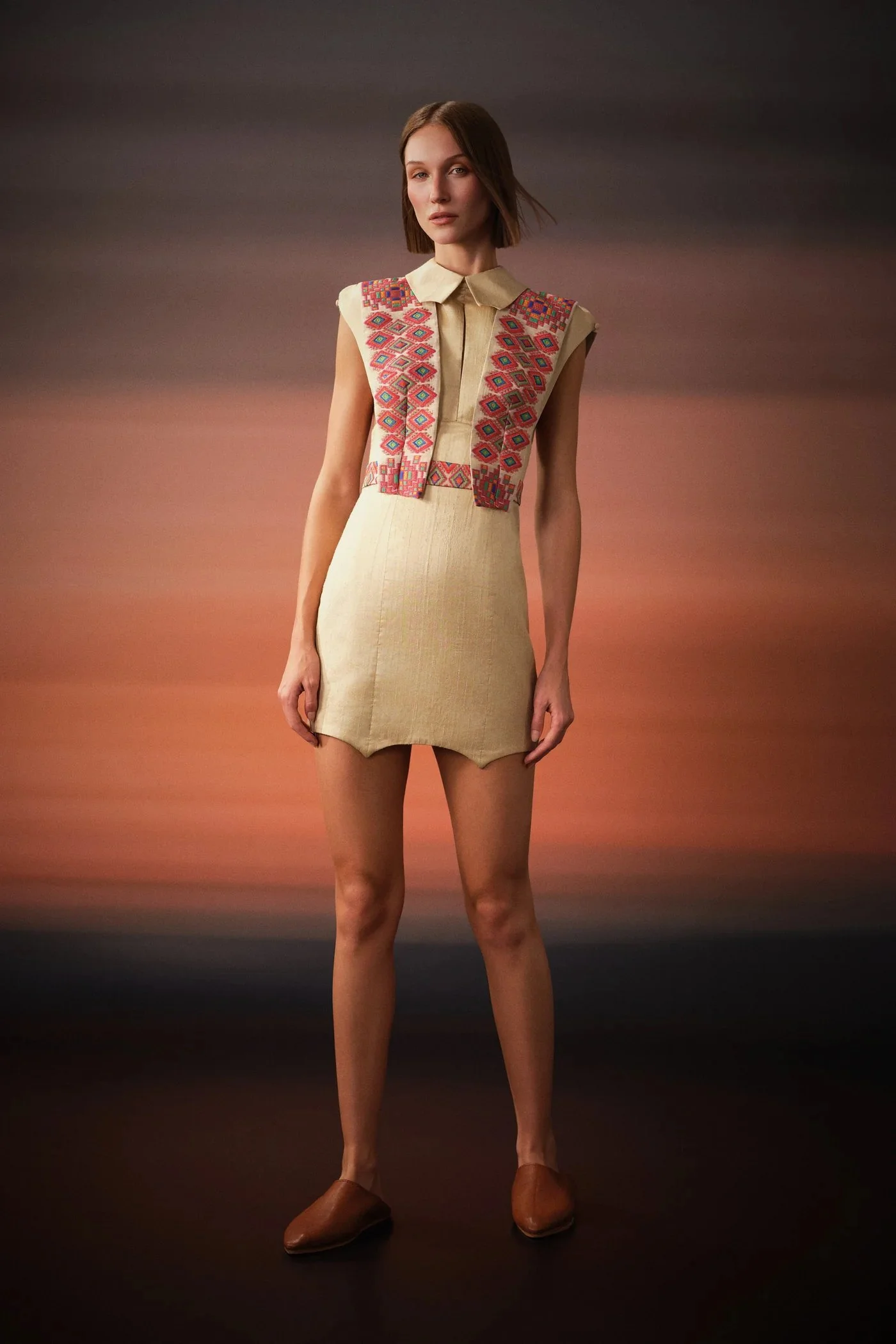 Model wearing a beige dress with embroidered panels on the top, standing against a sunset-colored background.