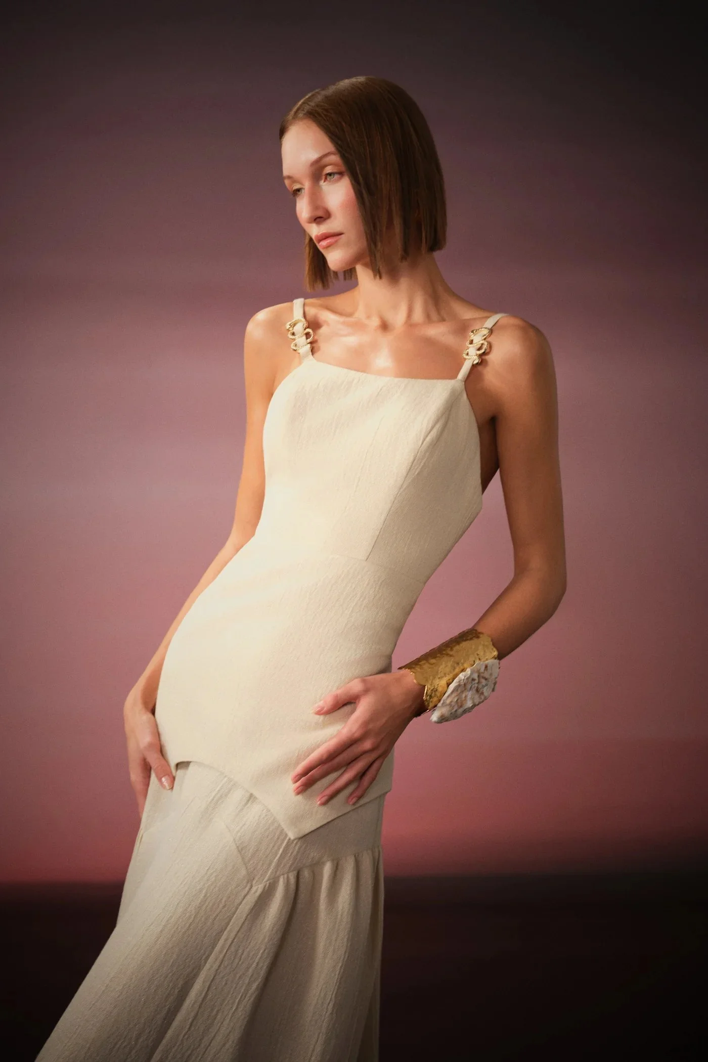 A woman in a cream-colored dress with decorative shoulder straps, standing against a gradient background. She has short, straight brown hair and is wearing a large cuff bracelet on her right wrist.