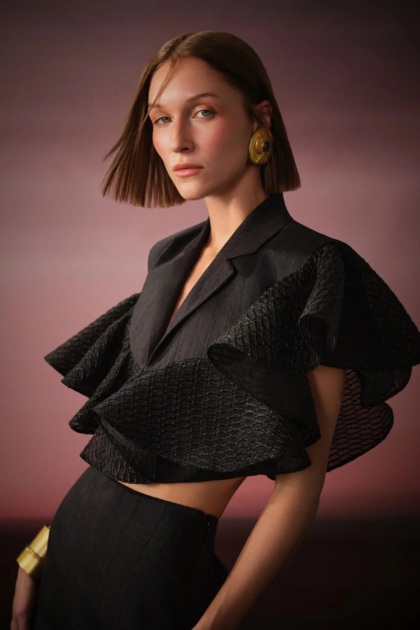 A woman with shoulder-length brown hair wearing a black textured blouse with ruffled sleeves, black pants, gold earrings, and a gold bracelet against a pinkish-purple background.