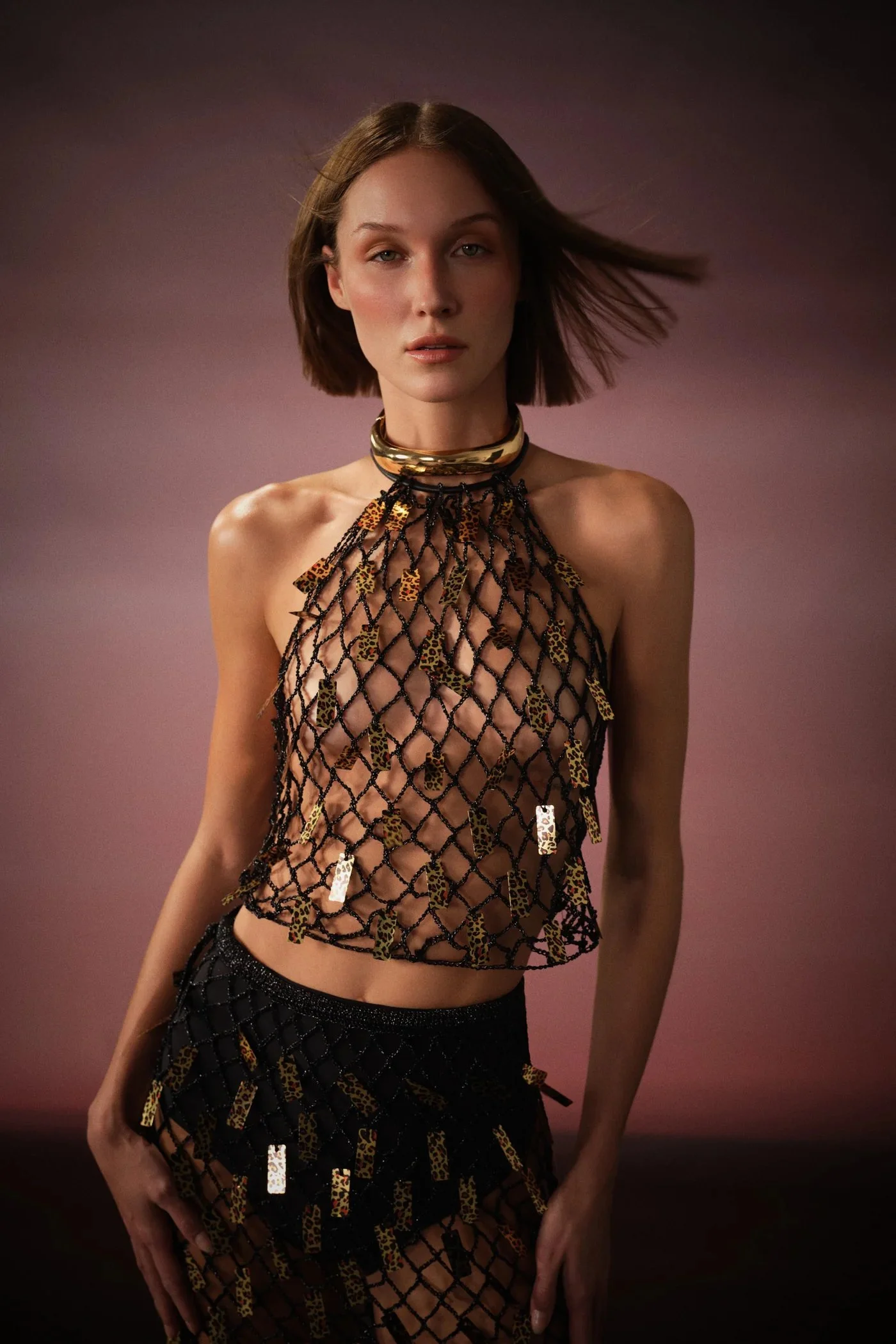 A woman with medium-length hair and fair skin posing against a pink background, wearing a black fishnet top with leopard-print accents and matching skirt, accessorized with a gold choker and necklace.