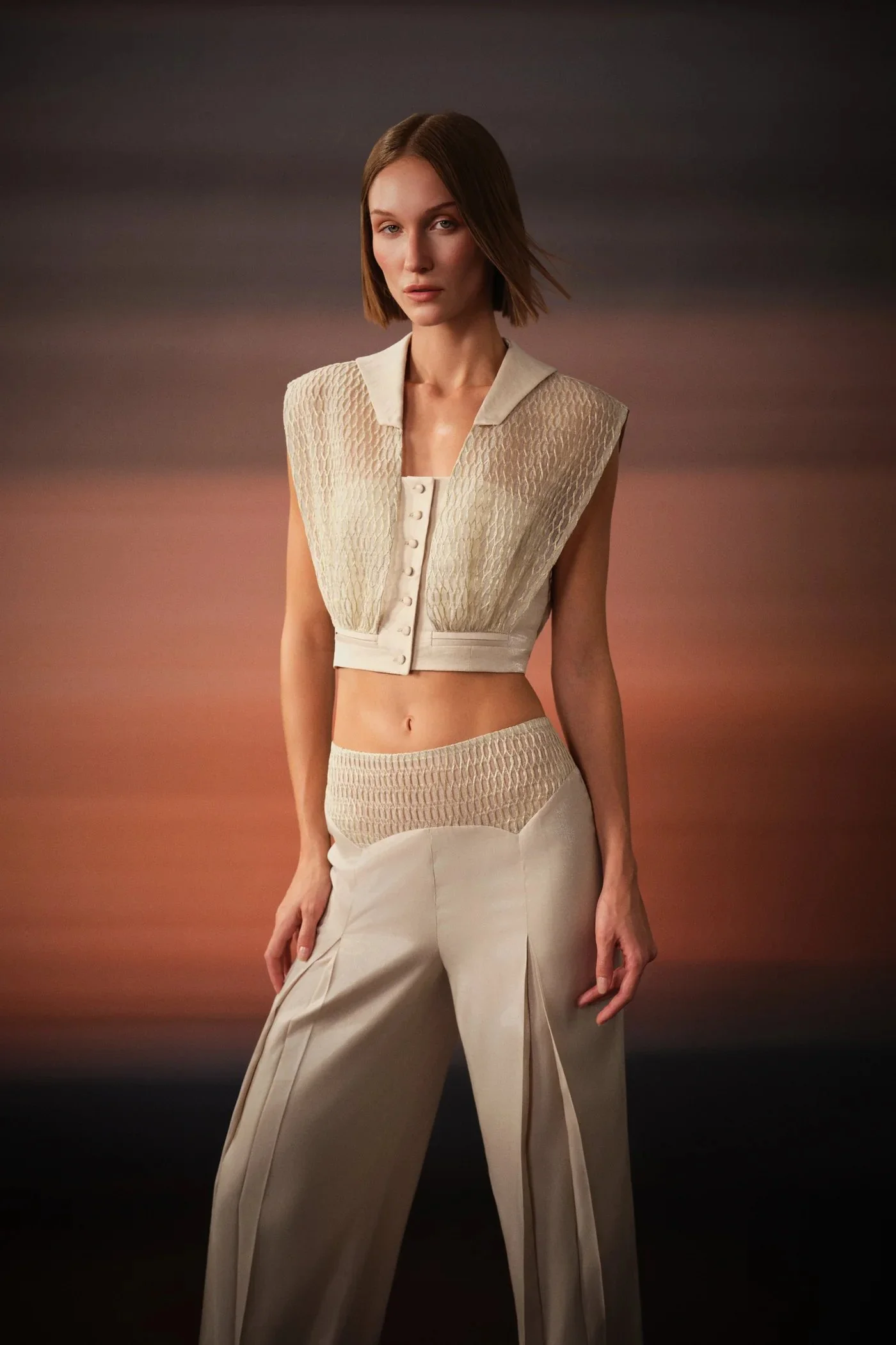 A woman with short brown hair wearing a cream-colored sleeveless knitted top with a collar and front buttons, and matching high-waisted wide-leg pants, standing against a blurred sunset background.