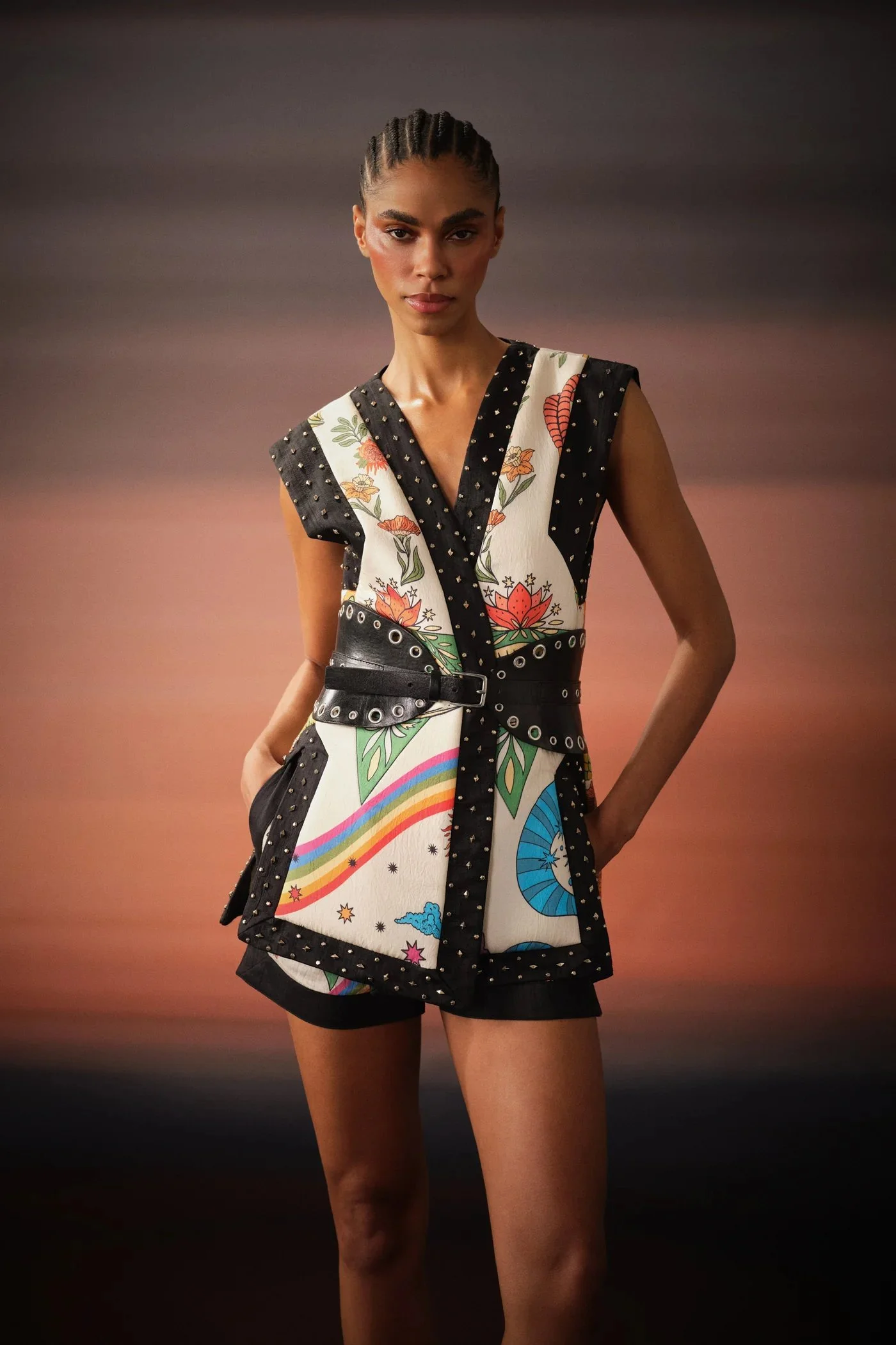 A woman posing against a sunset background, wearing a sleeveless, colorful, patterned vest with black trim and a matching belt, and black shorts.