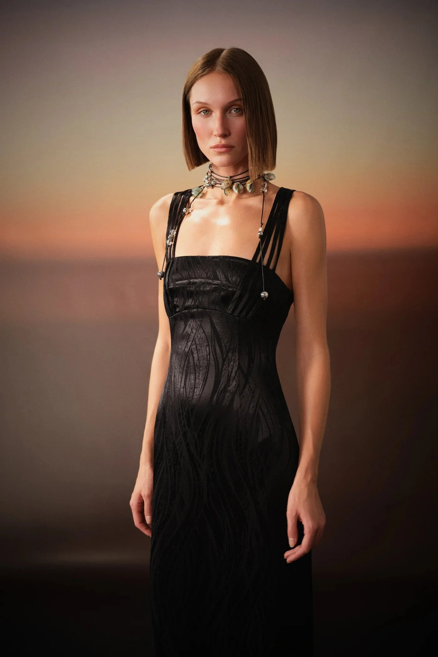 Young woman in a black dress standing in front of a sunset background.