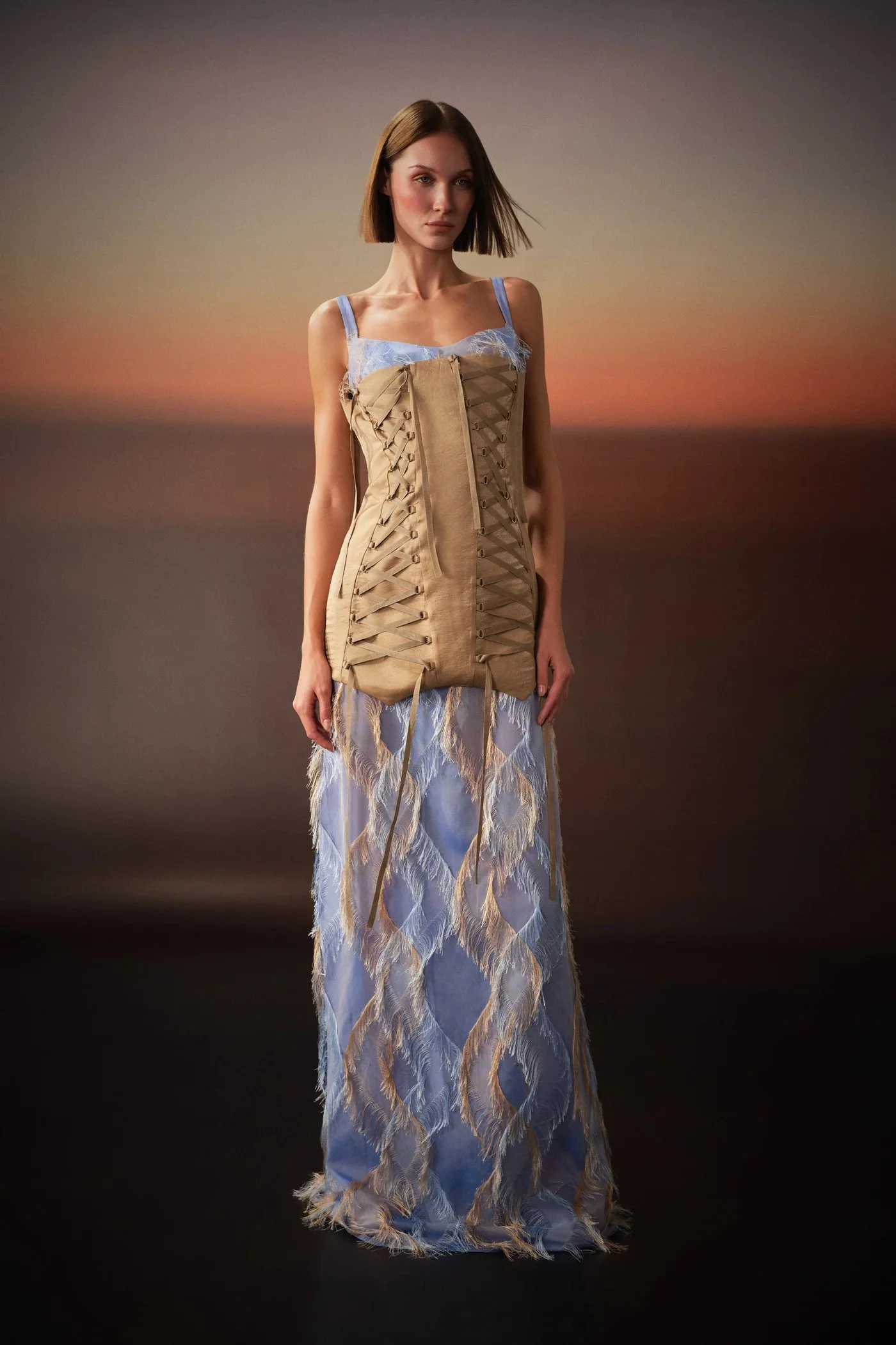 A woman standing outdoors during sunset, wearing a dress with feather-like patterns and a lace top under a beige dress with laced sides.