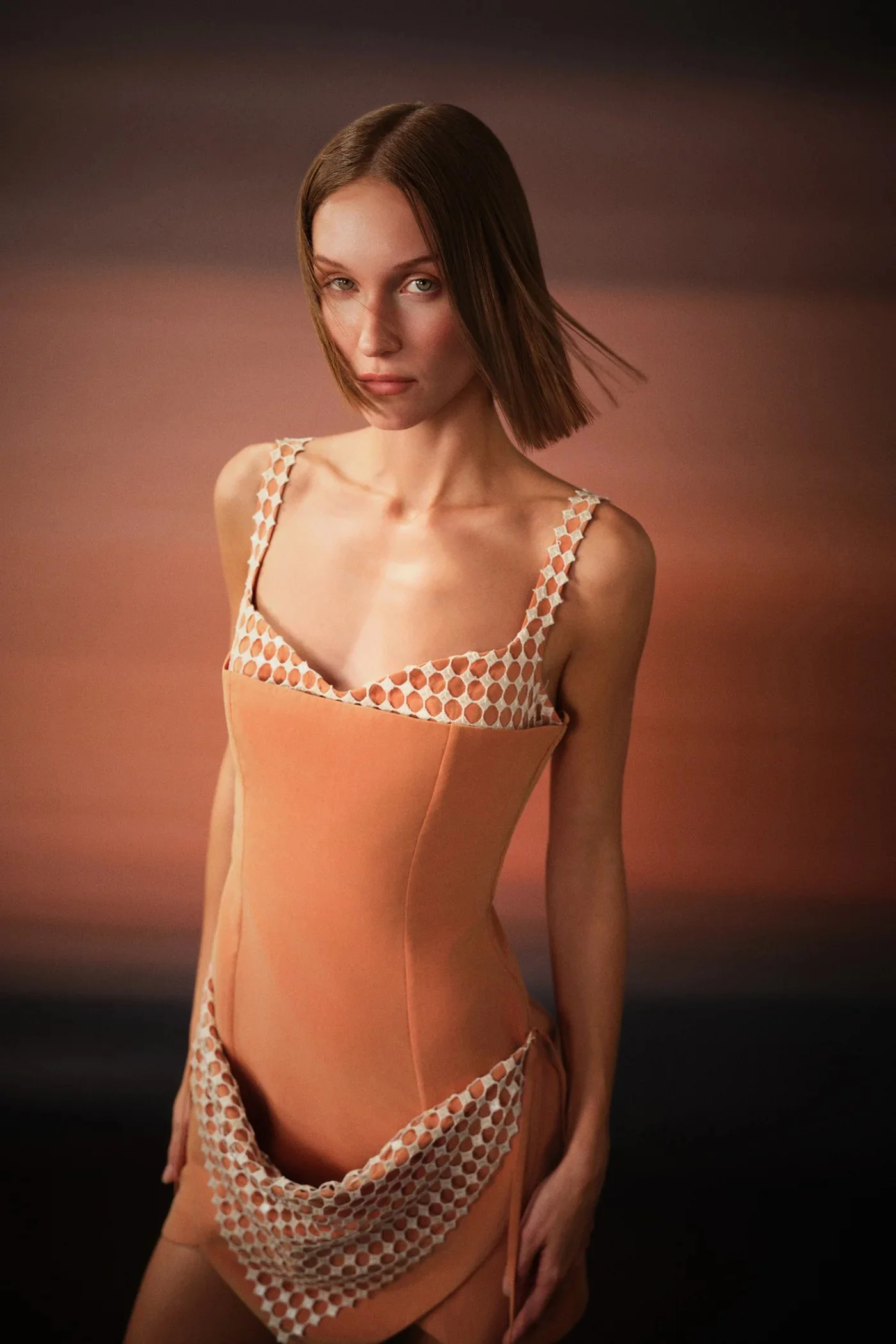 A young woman with medium-length brown hair and fair skin stares at the camera against a softly blurred pink and brown background, wearing a peach-colored dress with mesh and polka dot details.