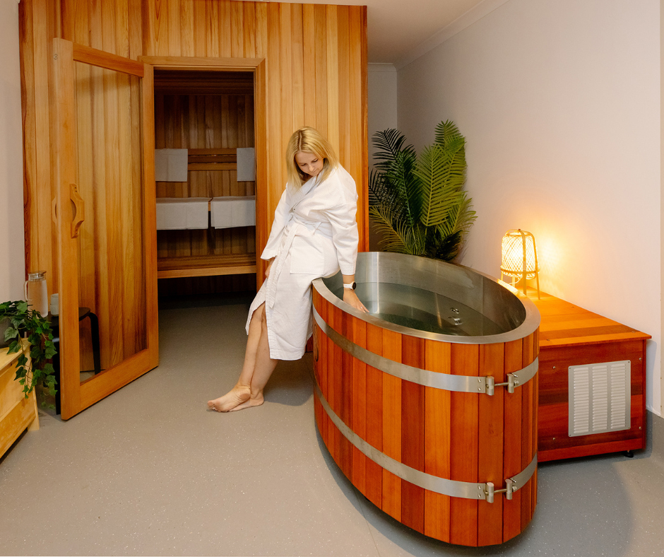 A woman in a white bathrobe soaking her feet in a wooden foot spa in a room with wooden and white walls, indoor plants, a bedside lamp, and a sauna room in the background.