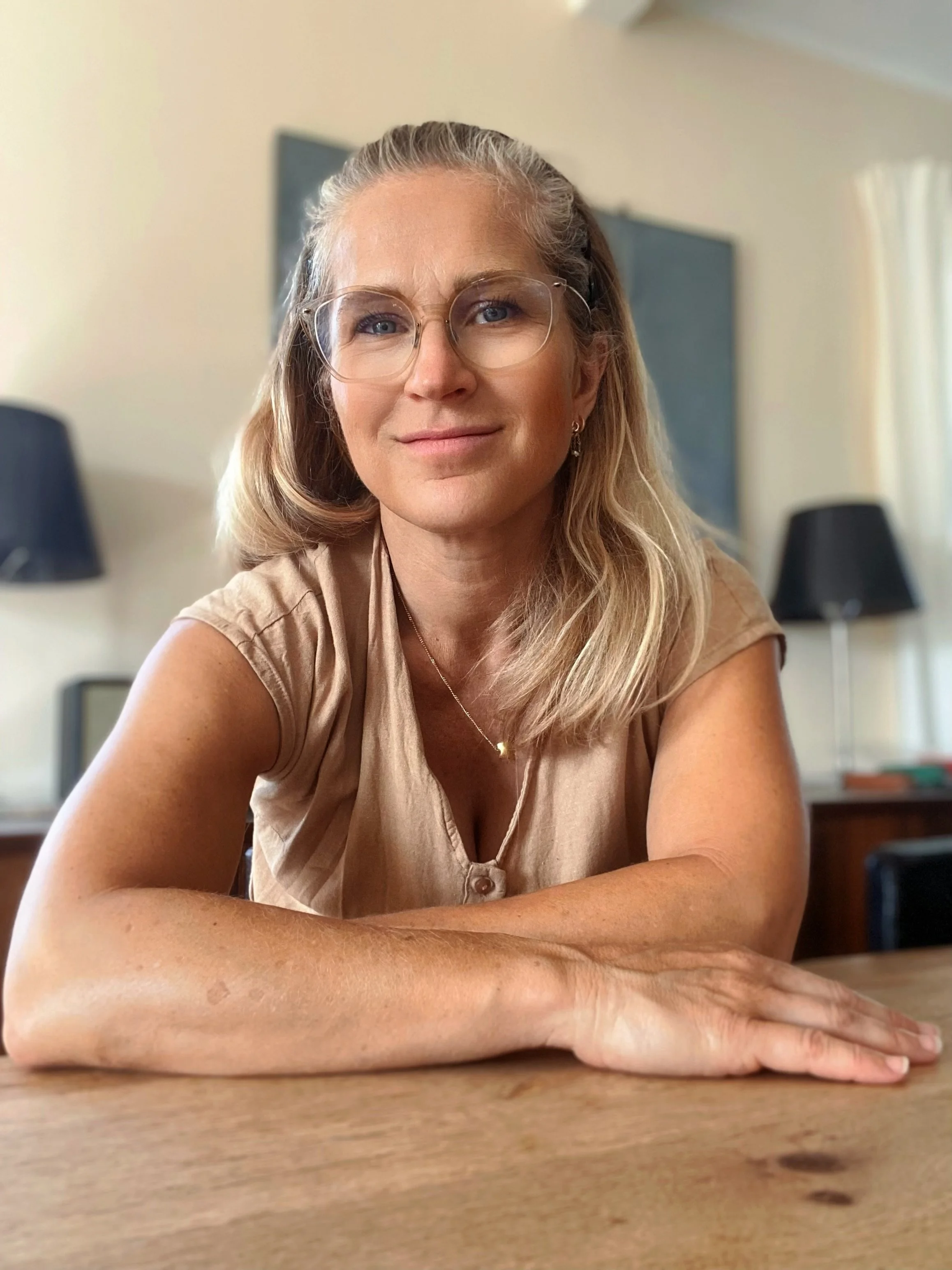Kvinde med briller og langt blondt hår sidder ved et træbord i et lyst rum, iført en beigefarvet bluse.