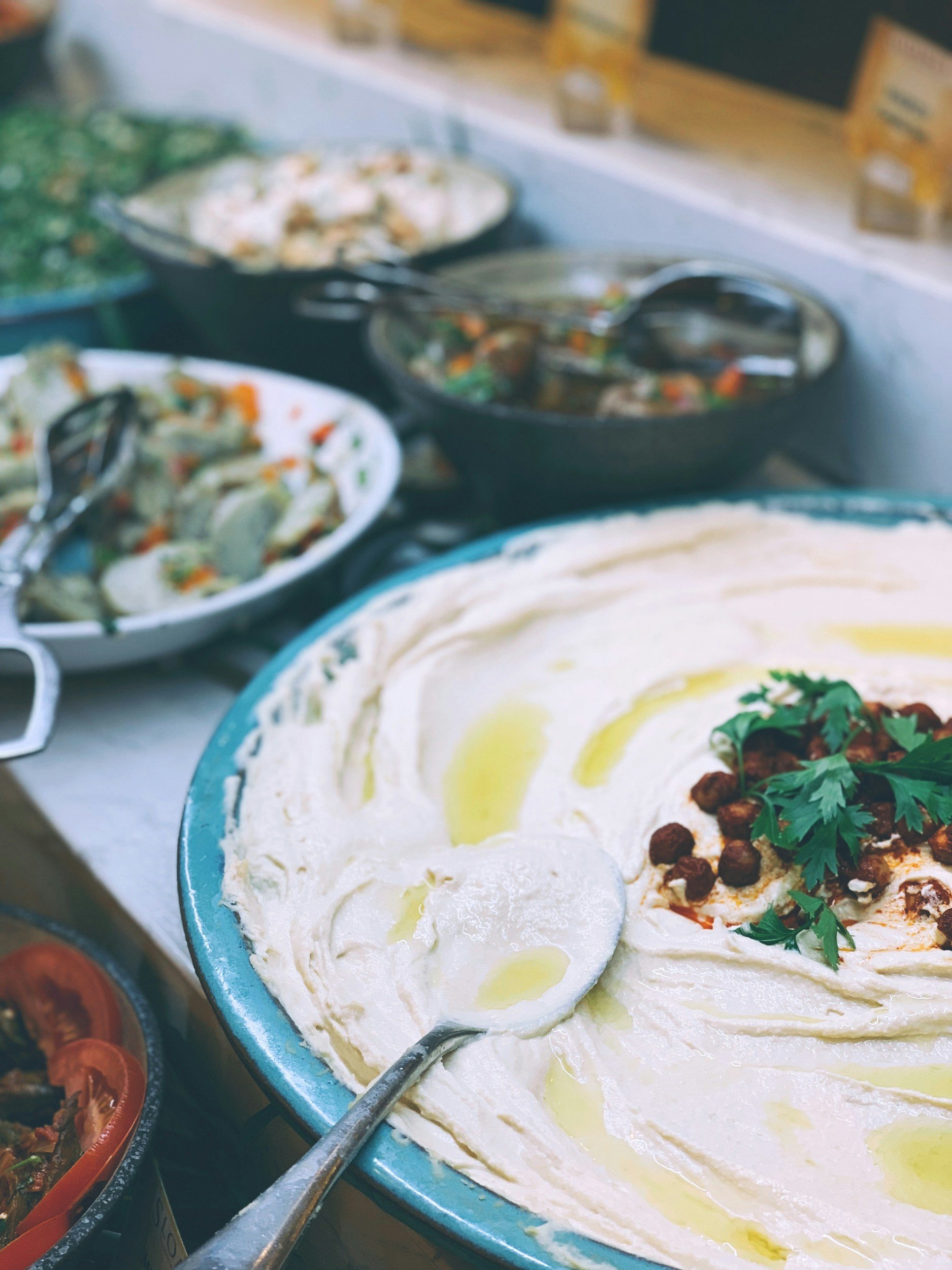 A large bowl of hummus topped with parsley, chickpeas, and olive oil. Several other bowls of salads and side dishes are visible in the background.
