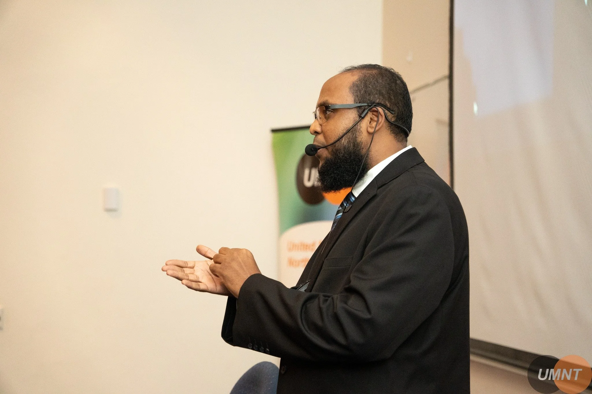 A man in a black suit giving a presentation in a classroom or conference room, with a UMNT banner in the background. Imam Mohanad Karmalah