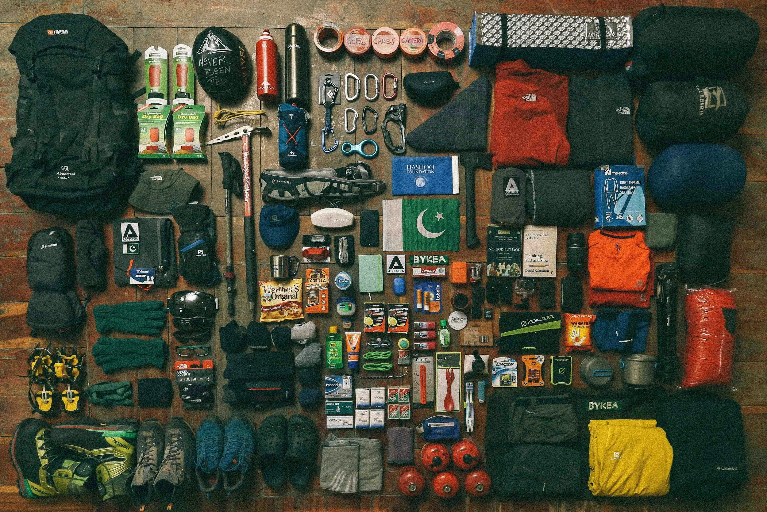 An organized collection of camping and outdoor gear laid out on a wooden floor, including backpacks, water bottles, tools, clothing, shoes, and various supplies.