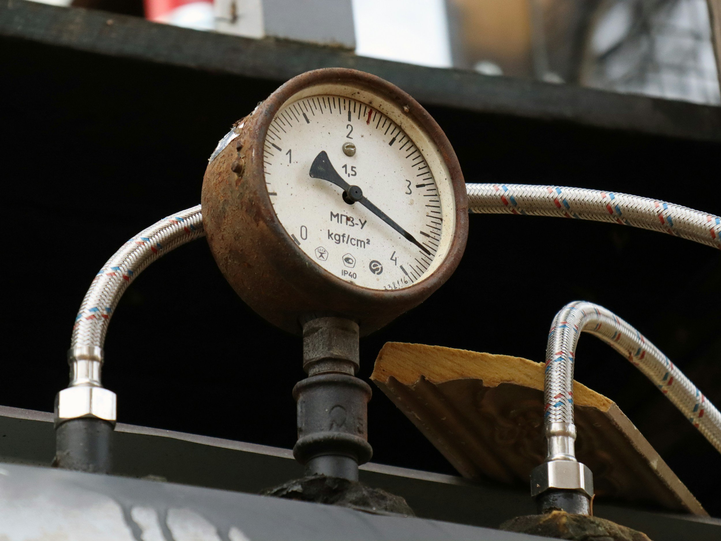 A pressure gauge attached to pipes with braided metal hoses, rusted on the exterior, showing pressure slightly below 1.5 kgf/cm².