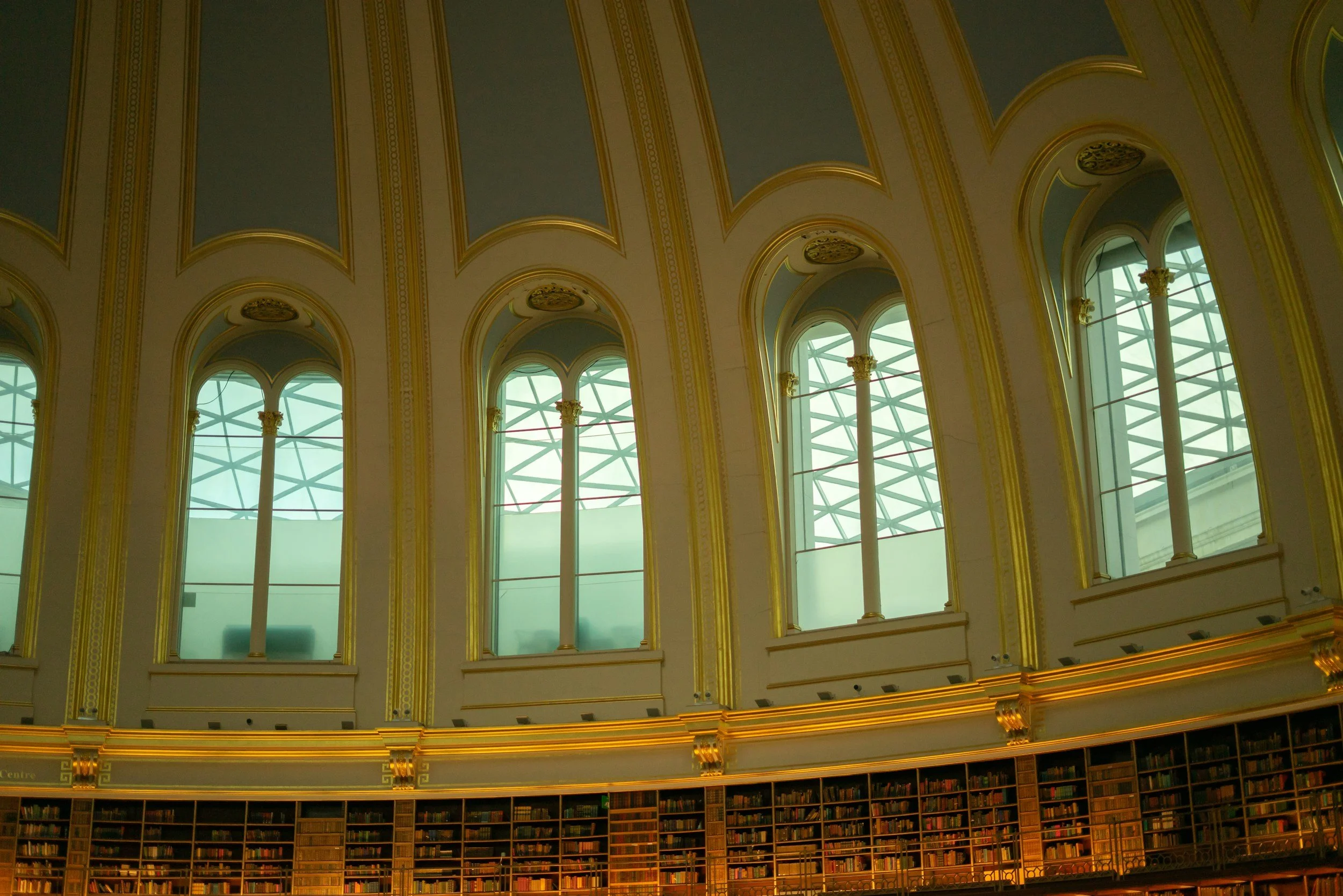 Interior of a grand library with tall arched windows and a domed ceiling, adorned with decorative gold accents and a curved bookshelf.