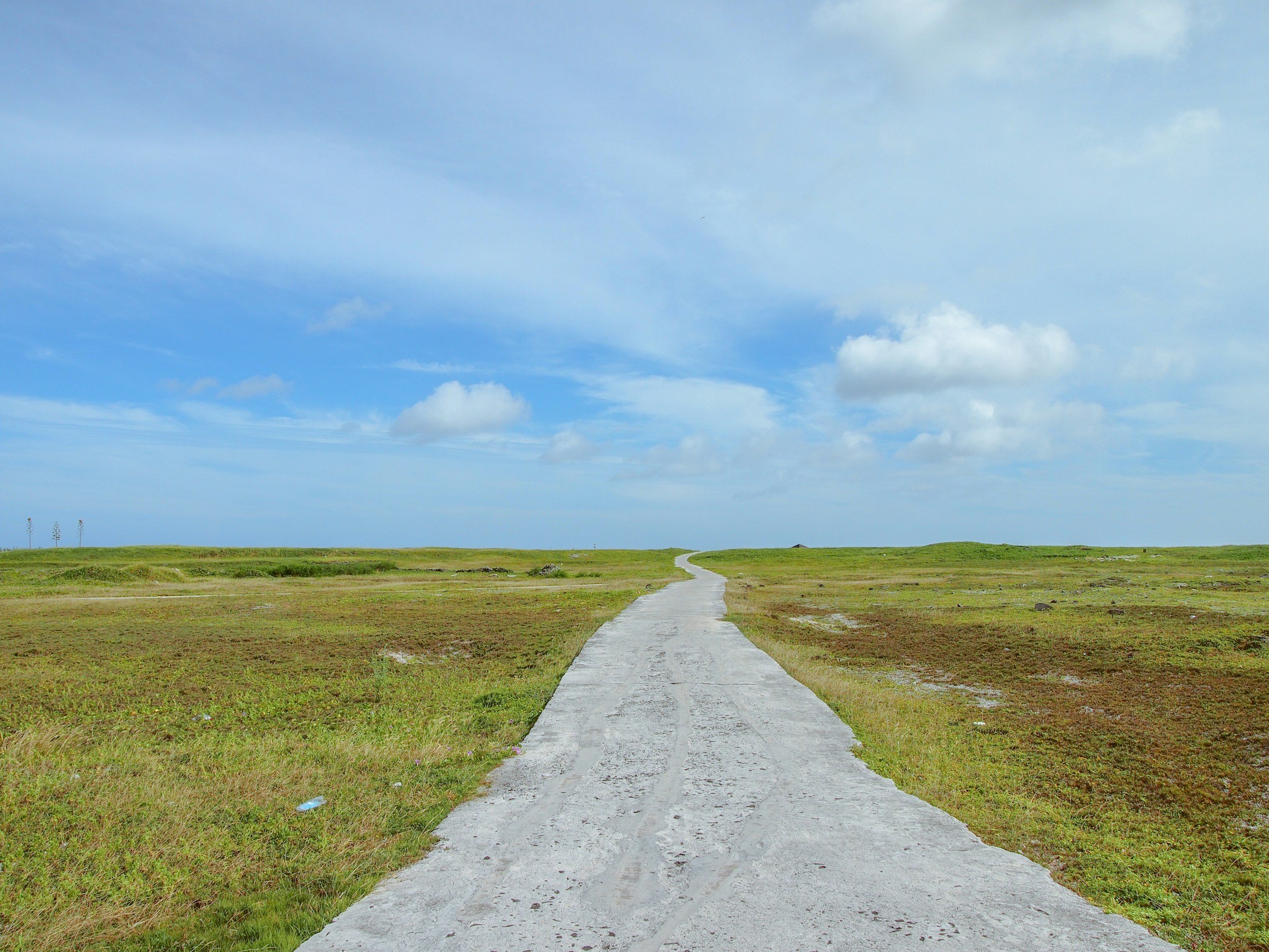 A long, narrow concrete road extends through a flat, grassy landscape toward the horizon under a partly cloudy sky.