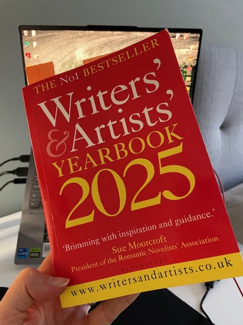 A photo of the front cover of the Writers' and Artists' Yearbook 2025. Helen holds the book up in front of a laptop on a desk.
