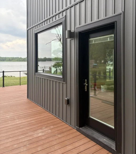 Exterior view of a black trimmed house with a large window and glass door and an AZEK deck with handrail, overlooking a lake with trees in the background.